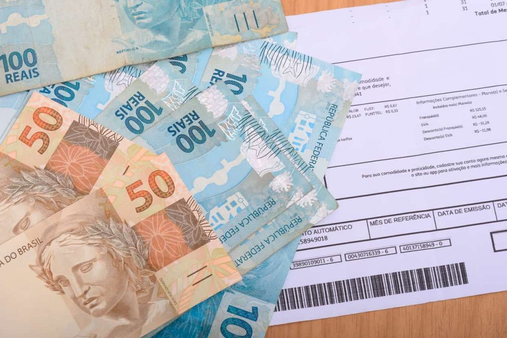 Diversas cédulas de 100 e 50 reais brasileiros espalhadas sobre uma conta de consumo impressa com código de barras visível. A imagem ilustra o pagamento de boletos e o custo de vida.